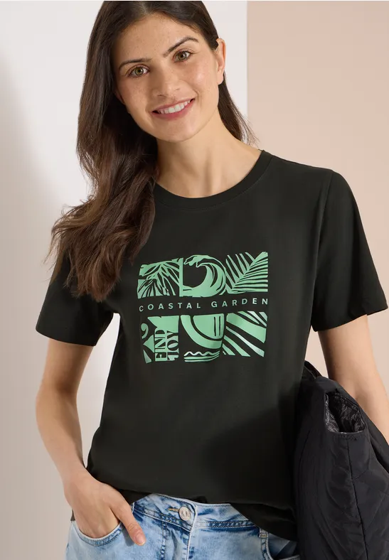 Vrouw met zwart T-shirt met groen plantenprint, lichte spijkerbroek en zwarte schoudertas.