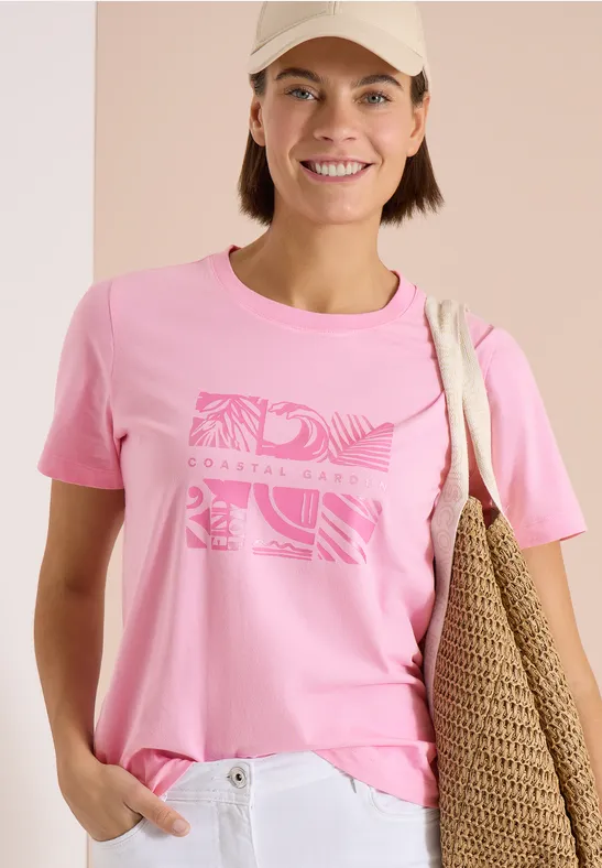 Persoon met roze t-shirt met print, een witte broek en een strooien schoudertas.