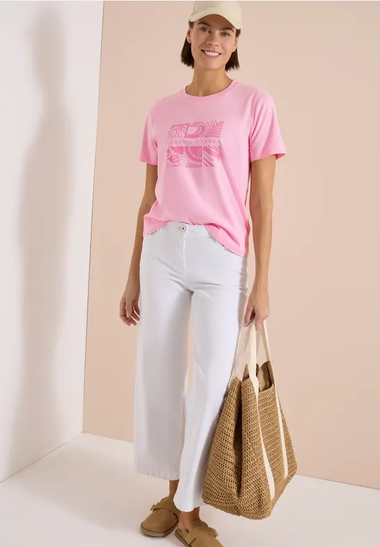 Persoon draagt roze T-shirt met print, witte wijd uitlopende broek, bruine loafers en een gevlochten tas.