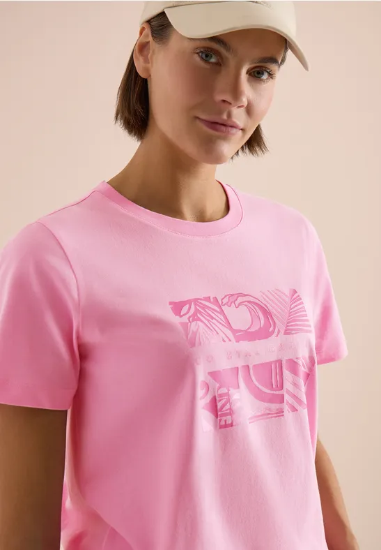 Een persoon draagt een roze T-shirt met abstract patroon en een pet.