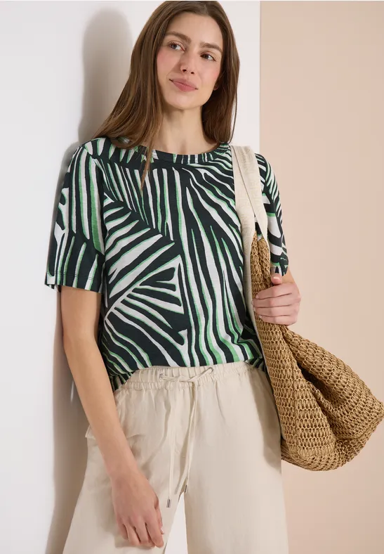 Frau trägt kurzärmliges Shirt mit grün-schwarzem Zebra-Muster, helle Hose und geflochtene Strohtasche.