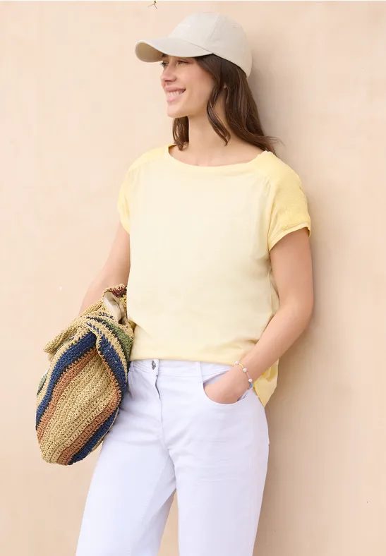 Frau steht vor beiger Wand, trägt gelbes T-Shirt, weiße Hose, Cap und bunte Häkel-Tasche.