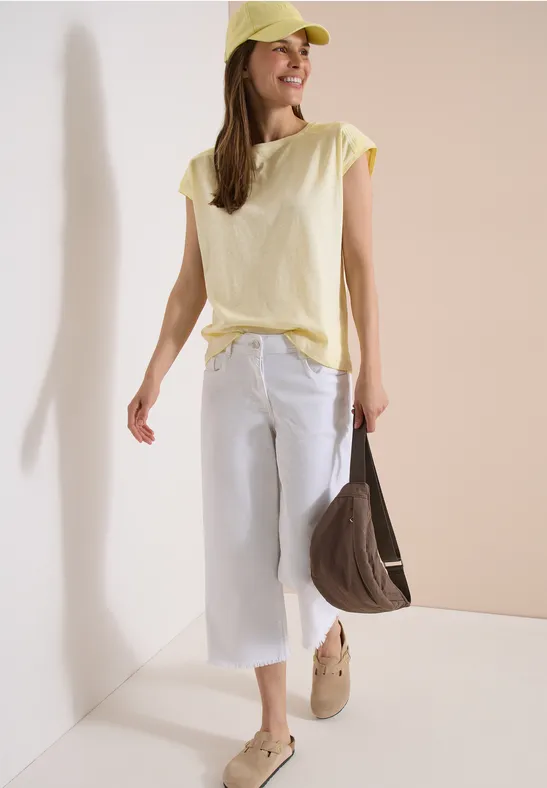 Frau in gelbem T-Shirt, weißer Culotte, gelber Mütze und brauner Tasche vor neutralem Hintergrund.