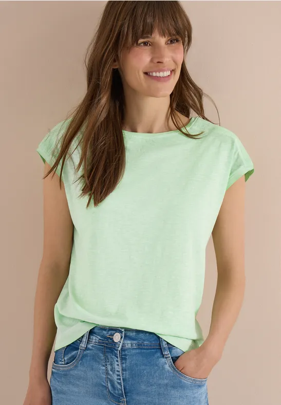 Femme portant un t-shirt vert menthe et un jean bleu, une main dans la poche, fond beige.