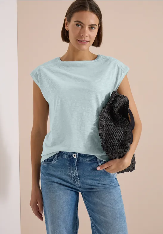 Frau trägt hellblaues ärmelloses Oberteil, blaue Jeans und hält schwarze Tasche.