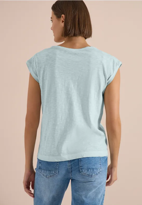 Eine Frau von hinten in hellgrauem T-Shirt und blauer Jeans vor hellem Hintergrund.