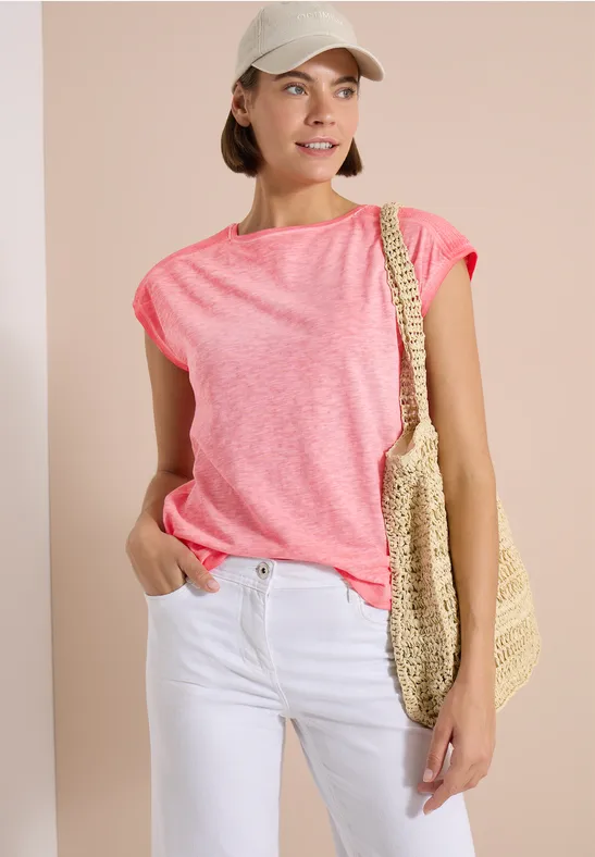 Frau im pinken T-Shirt und weißen Jeans mit Strohtasche und Baseballcap vor hellem Hintergrund.