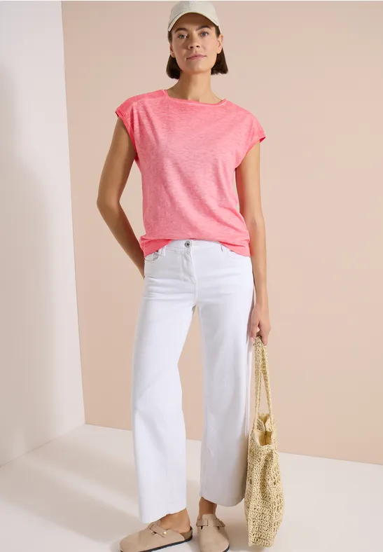 Frau trägt pinkes T-Shirt, weiße gerade Hose und Basttasche vor hellem Hintergrund.