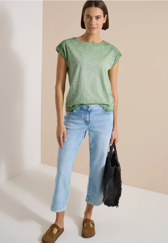 Frau trägt grünes T-Shirt, helle gerade Jeans, braune Wildleder-Slipper und schwarze Netz-Tasche.