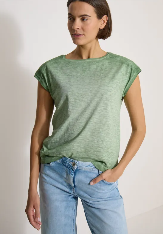 Eine Frau mit lockerem grünen T-Shirt und blauen Jeans, eine Hand in der Hosentasche.
