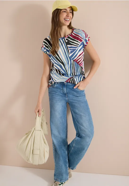 Person trägt bunte gestreifte Bluse, weite Jeans, gelbe Cap und große beige Schultertasche.