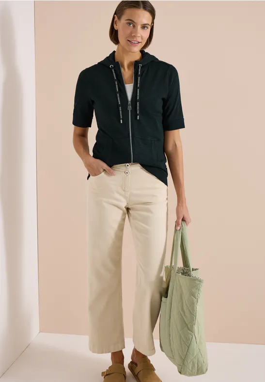 Frau in schwarzem Kurzarm-Zipshirt, hellem Culotte-Hose und gestepptem grünen Shopper