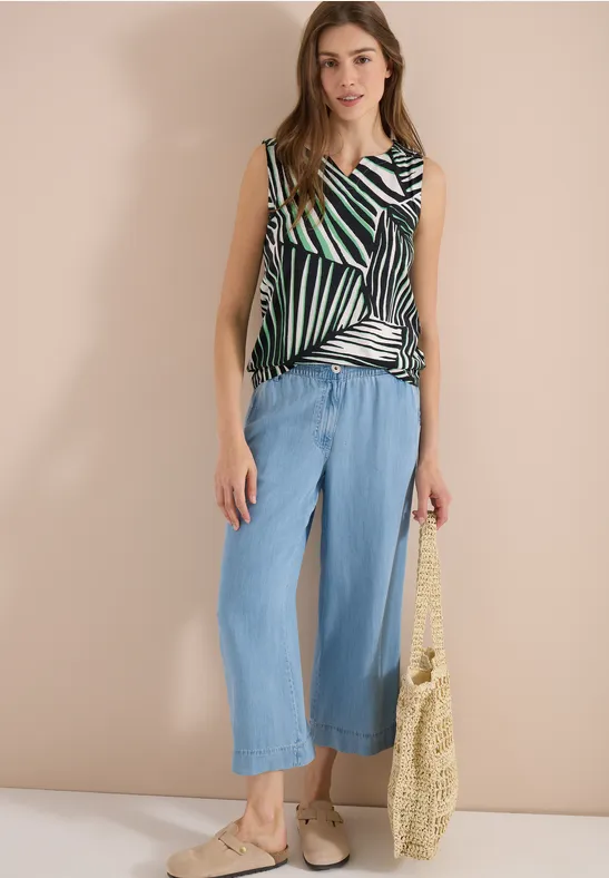 Frau trägt gestreiftes Top, weite blaue Culotte und Basttasche vor neutralem Hintergrund.