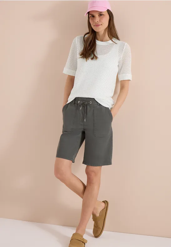 Vrouw staat voor een beige achtergrond, draagt wit T-shirt, grijze bermudashort en bruine loafers.