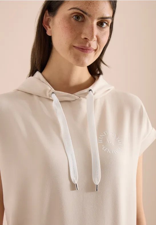 Person mit langen dunklen Haaren trägt beiges ärmelloses Kapuzenshirt mit Kapuze und weißen Kordeln.