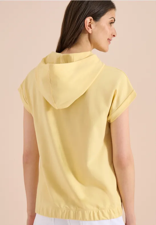 Frau von hinten in hellgelbem, kurzärmeligem Kapuzenoberteil.