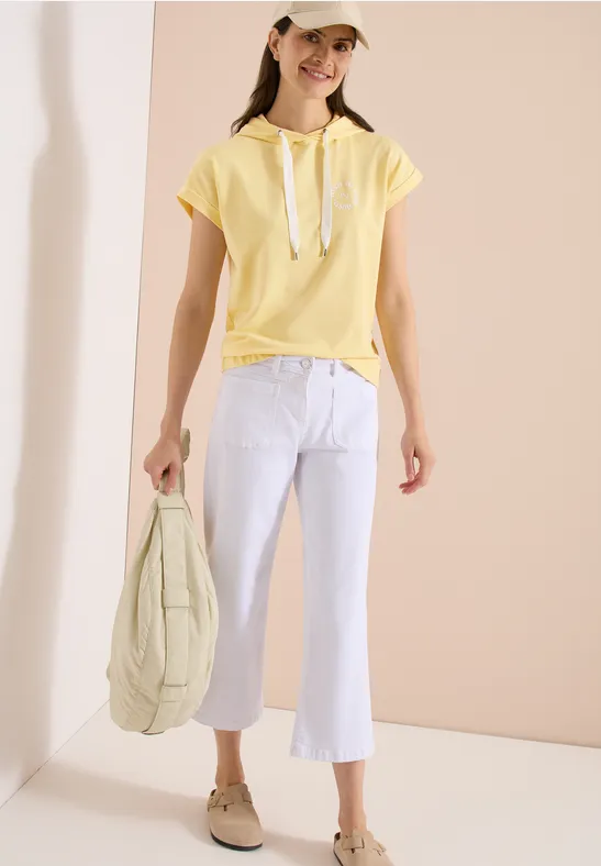 Junge Frau in gelbem Kapuzen-Shirt, weißer Schlaghose, beiger Kappe und Slippern hält gesteppte Tasche.