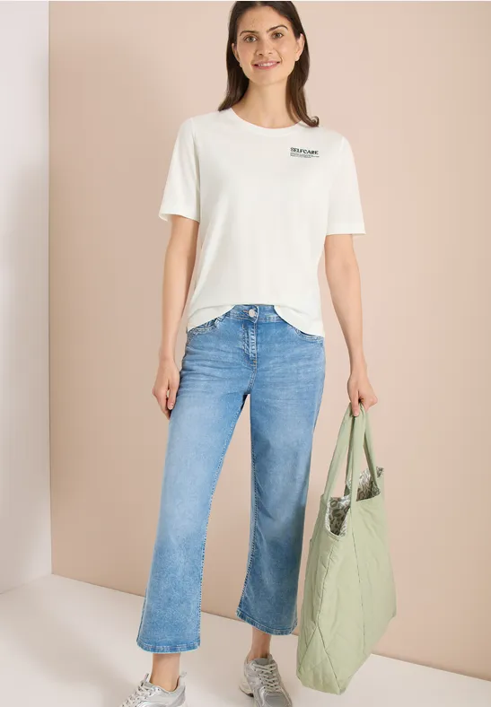 Frau in weißem T-Shirt, blauen Jeans und großer grüner Tasche vor neutralem Hintergrund.