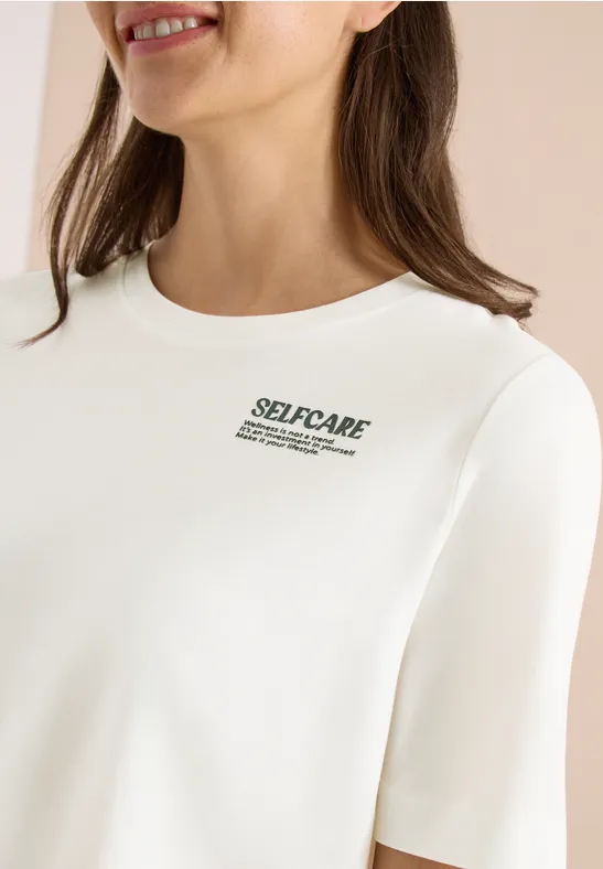 Eine Frau trägt ein weißes Shirt mit kleinem 'SELF-CARE'-Aufdruck.