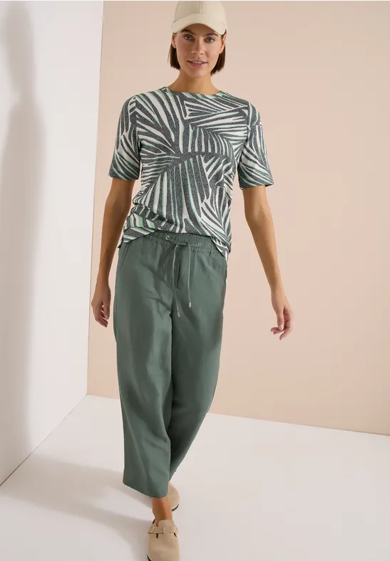 Vrouw in groen t-shirt met bladprint en wijde groene broek tegen neutrale achtergrond.