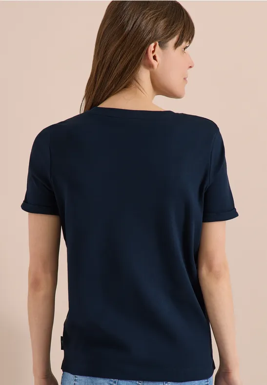 Frau von hinten mit dunkelblauem Kurzarmshirt und langen braunen Haaren vor beigem Hintergrund.