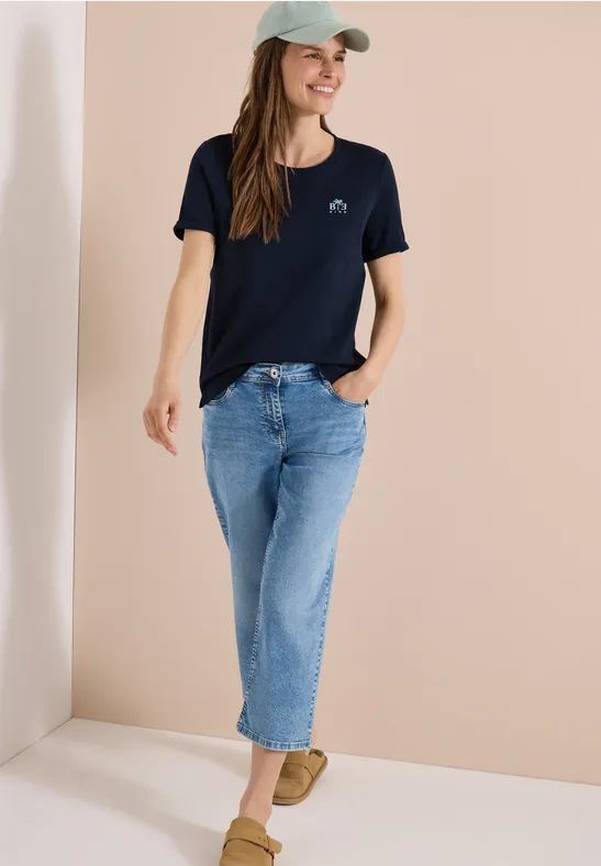 Eine Frau trägt dunkelblaues T-Shirt, blaue Jeans, beiges Slip-on und grüne Kappe vor beigem Hintergrund.