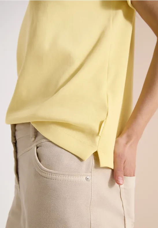 Seitliche Ansicht einer Person in beigen Jeans und lockerem gelbem Shirt vor hellem Hintergrund.