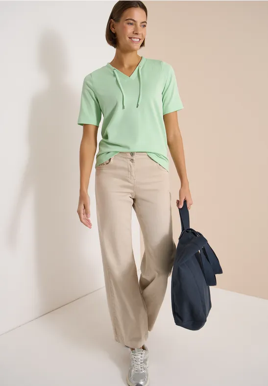 Vrouw in groen capuchon-T-shirt, beige wijd uitlopende broek en blauwe tas bij lichte muur.
