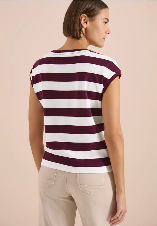 Eine Frau steht mit dem Rücken zur Kamera und trägt ein weiß-bordeauxgestreiftes T-Shirt mit beigen Hosen.