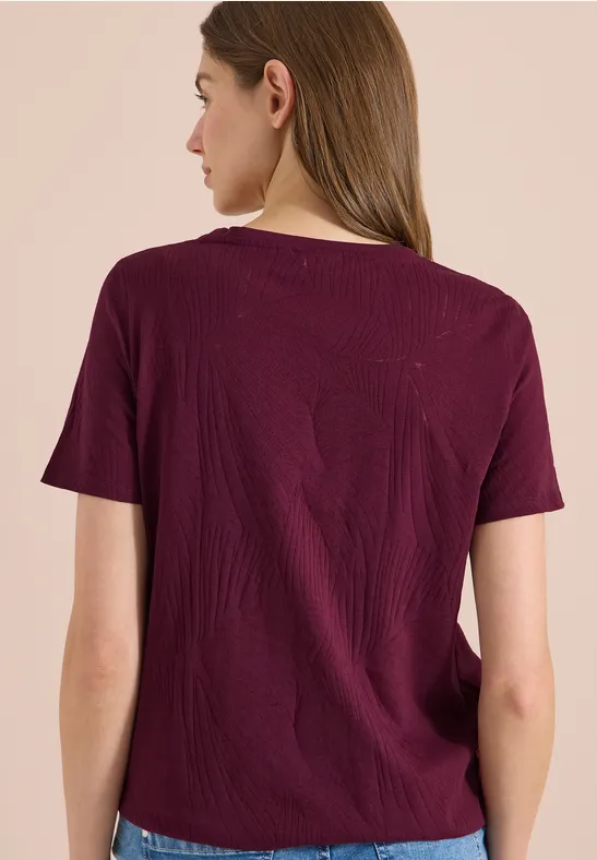 Femme vue de dos portant un T-shirt bordeaux sur fond neutre et clair.