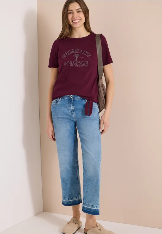 Femme en t-shirt bordeaux, jean clair et ballerines beiges, sac épaule, posant devant un mur beige.