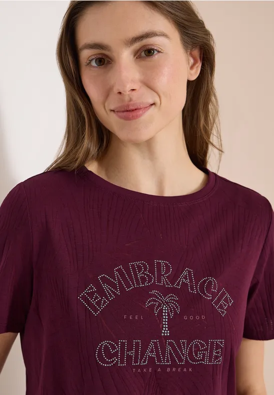Femme en t-shirt bordeaux avec l'inscription 'Embrace Change' et un motif palmier.