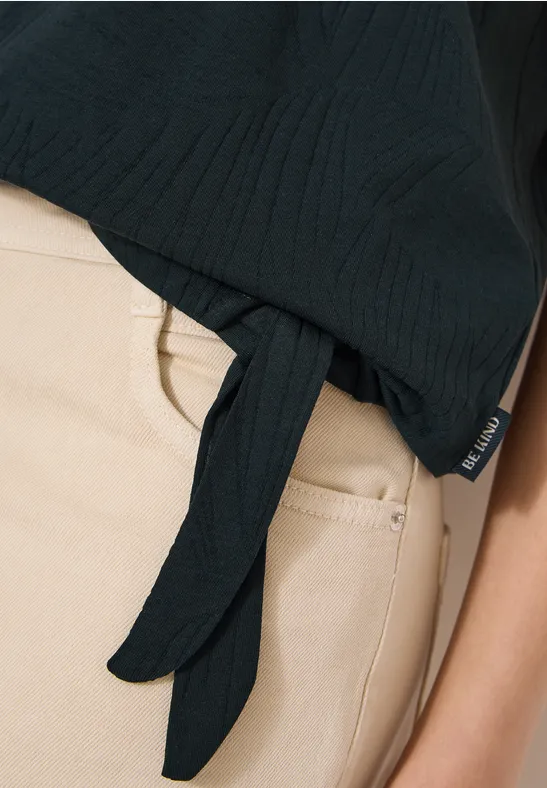 Nahaufnahme einer Person mit beiger Hose und schwarzem, seitlich geknotetem Oberteil.