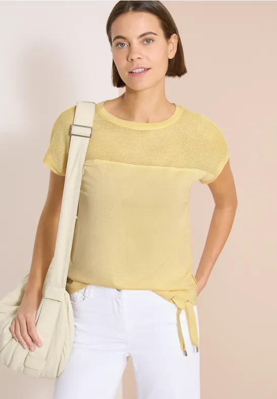 Vrouw met gele korte mouw top, witte broek en witte schoudertas.