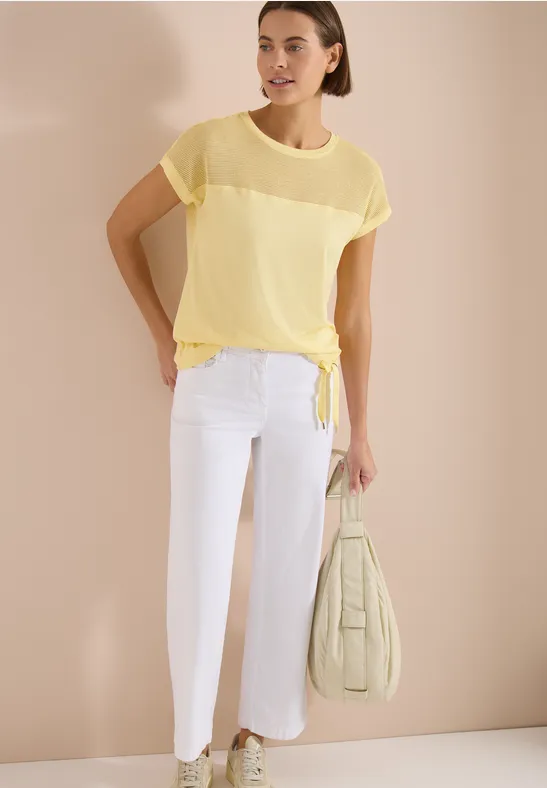 Model draagt transparant geel kort shirt met knoop, witte cropped broek en een pastelgroene tas.