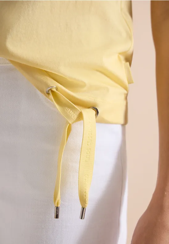 Geel T-shirt aan de taille geknoopt, gecombineerd met een witte rok en een arm in beeld.