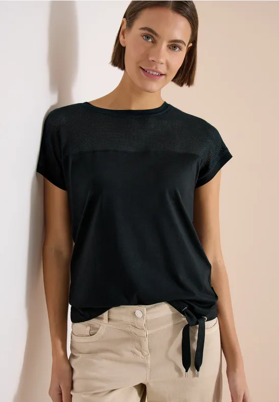 Vrouw leunt tegen muur, draagt zwart t-shirt met strik en lichte broek.