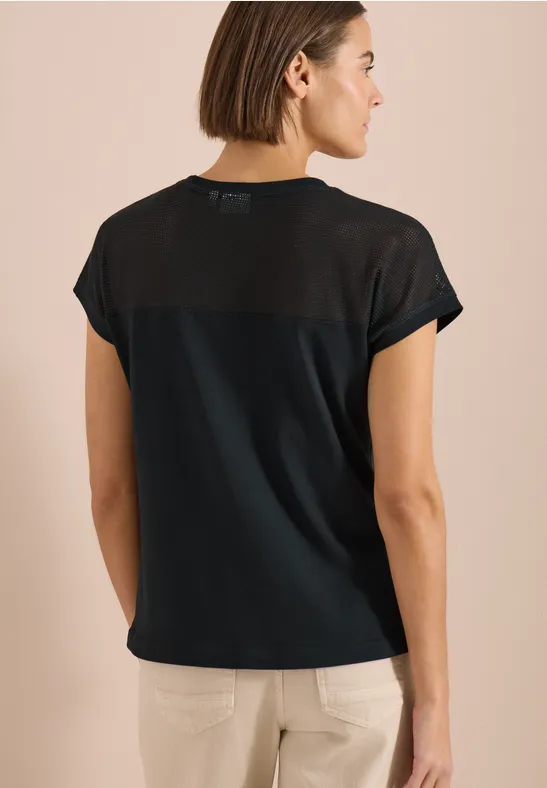 Achteraanzicht van een vrouw in een zwart T-shirt met transparante rugpartij en lichte broek.