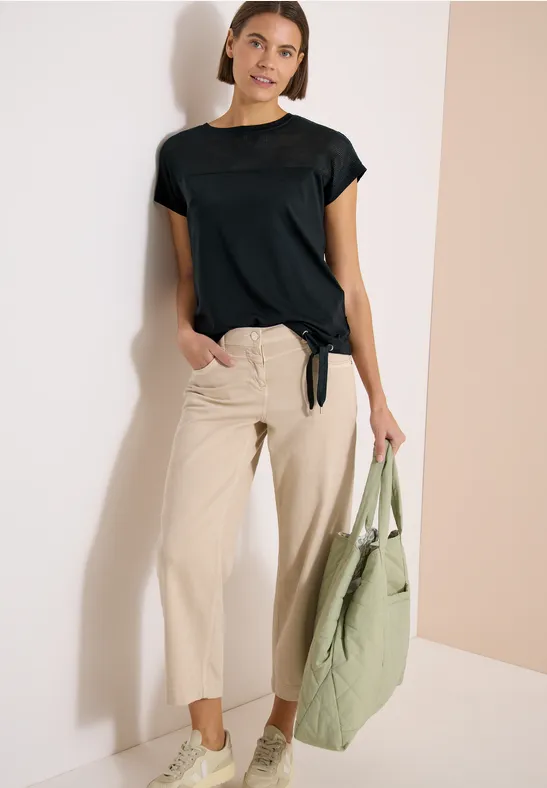 Model draagt een zwart t-shirt met korte mouwen, een beige broek en houdt een groene tas vast, leunend tegen een muur.
