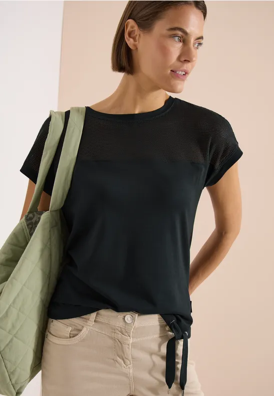 Vrouw draagt donkerblauw T-shirt met transparante inzet bij de schouders, knoop onderaan en groene tas.