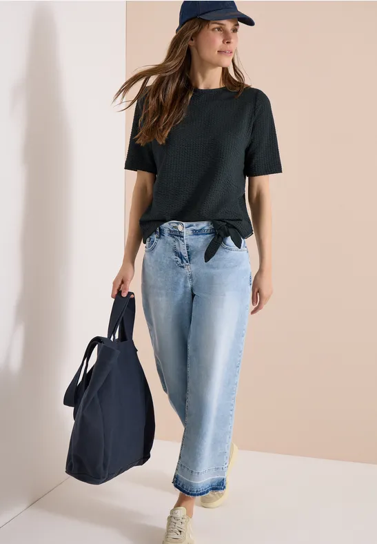 Vrouw in zwart T-shirt, spijkerbroek en pet, met een grote blauwe tas.