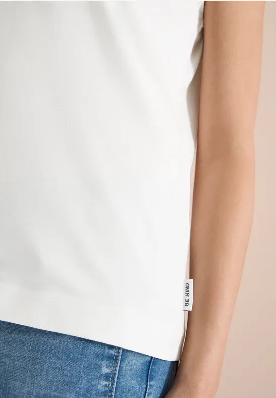 Weißes ärmelloses Oberteil aus leichtem Stoff, sitzt locker über blauen Jeans, mit seitlichem Label.