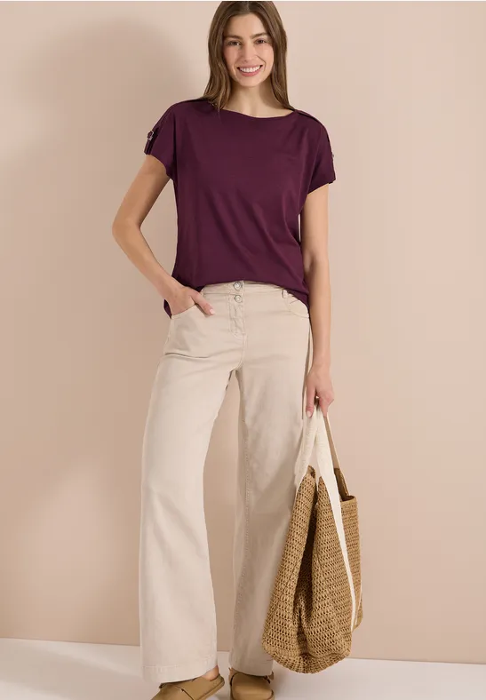 Frau trägt lila T-Shirt, weiße weite Hose und hält eine geflochtene Tasche.