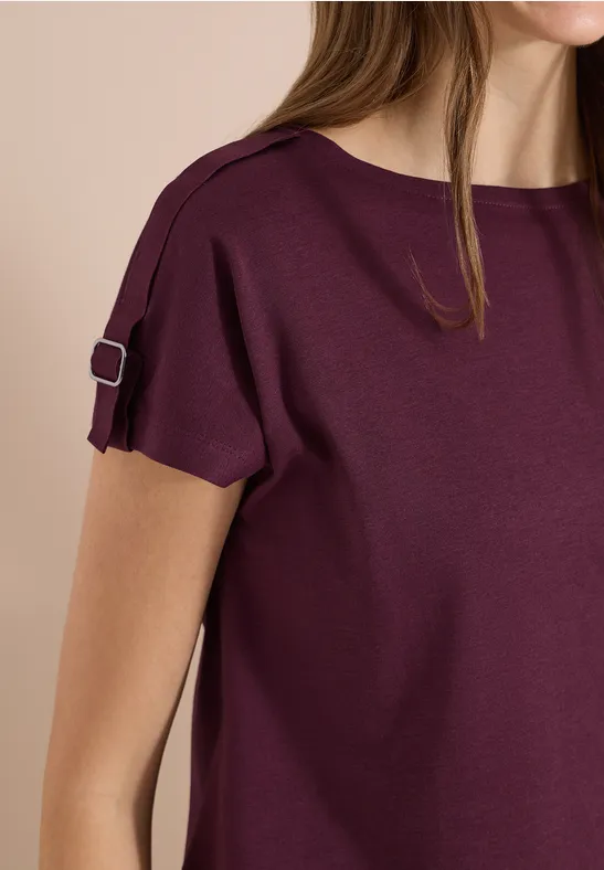 Eine Frau trägt ein kurzärmeliges, pflaumenfarbenes Oberteil mit einer kleinen Schlaufe und Schnalle am Ärmel.