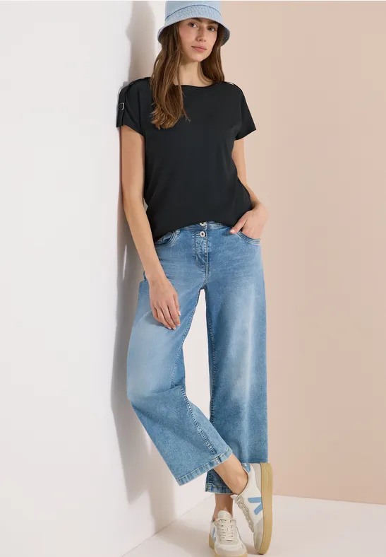 Frau in schwarzem T-Shirt, weiten blauen Jeans und weißen Sneakern vor zweifarbigem Hintergrund.