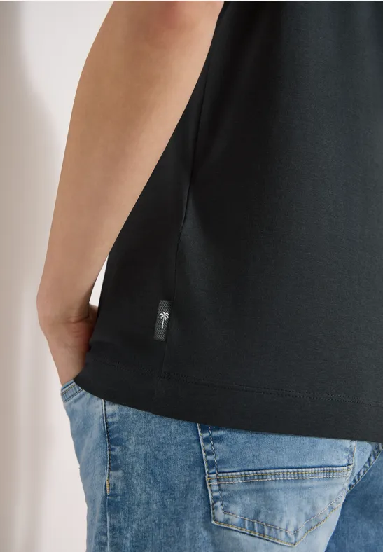 Eine Person trägt ein schwarzes T-Shirt und blaue Jeans, Hand in Hosentasche.