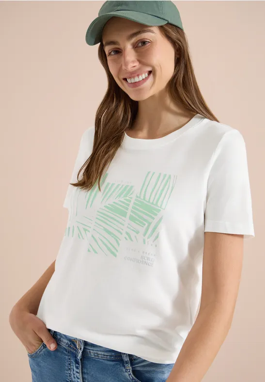 Femme en t-shirt blanc à motif de feuilles vertes, jean et casquette bleue sur fond neutre.