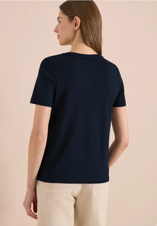 Frau steht seitlich mit Blick nach rechts, trägt ein dunkelblaues T-Shirt, vor beigem Hintergrund.
