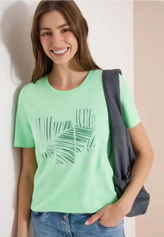 Frau in grünem T-Shirt mit Blattmuster, eine Hand in der Tasche, Rucksack über der Schulter.