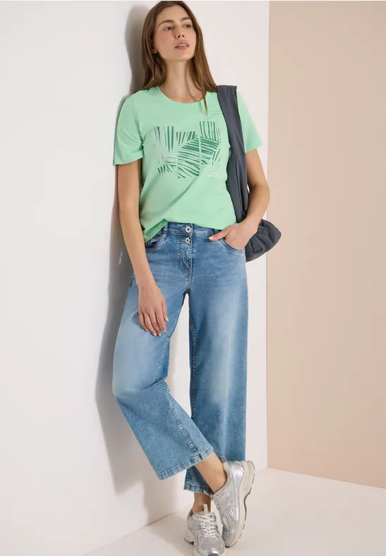Frau lehnt an Wand, trägt grünes T-Shirt mit Print, weite Jeans-Culotte, weiße Sneaker und Denim-Tasche.